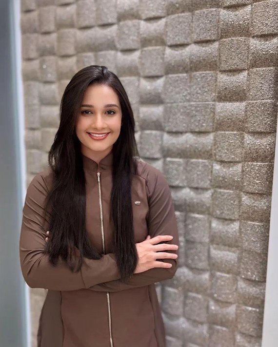 Dra. Anna Katharina, de jaleco marrom e braços cruzados, no consultório